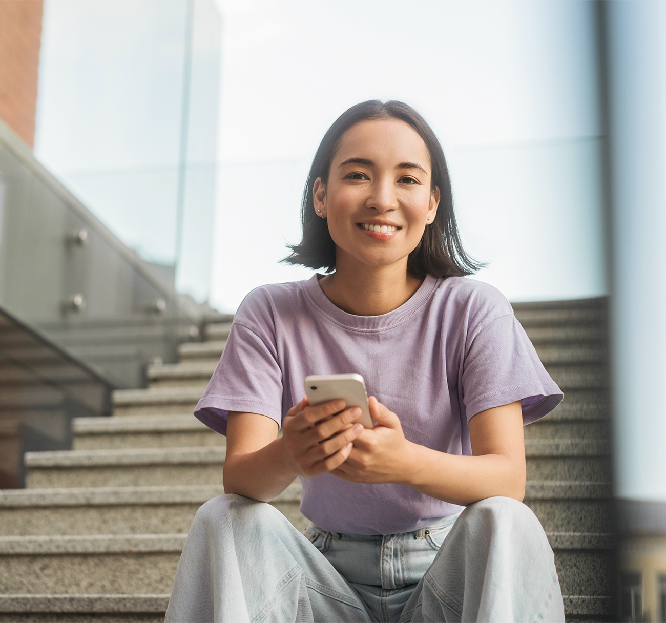 Smiling young woman sitting on steps with a phone, representing welcoming dental patient experience – dentist williamsville ny