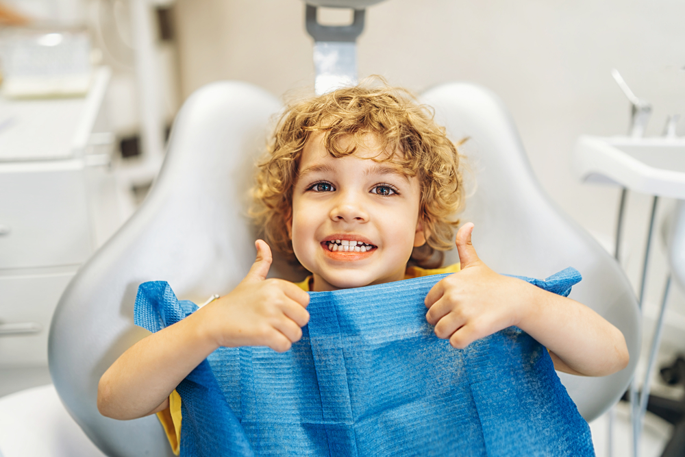 Smiling child sitting in a dental chair with thumbs up, showing a positive kids dental experience – pediatric dentist williamsville ny